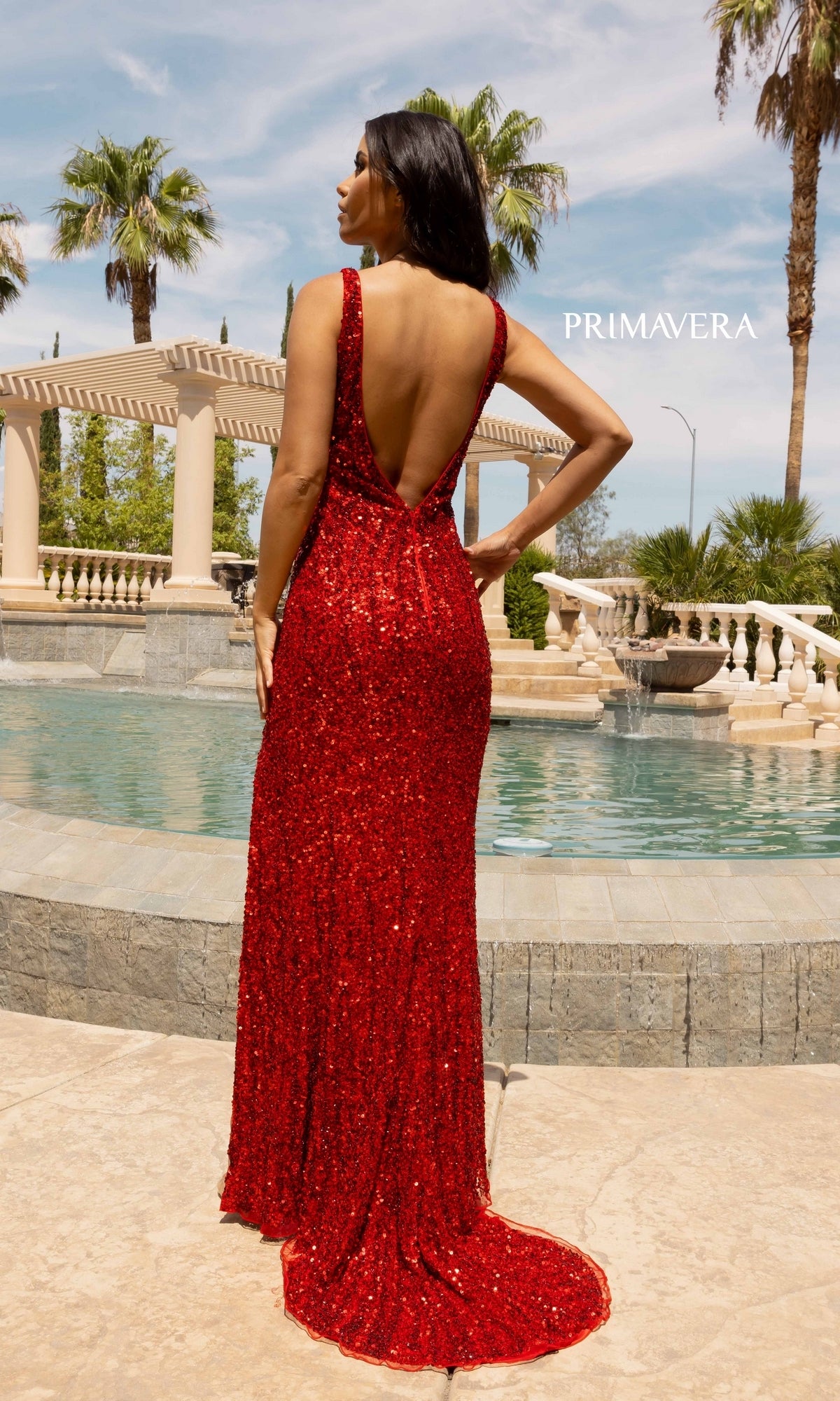 A woman wearing the Open-Back Long Sequin Prom Dress 3912 stands by a fountain, facing away, with palm trees and a pergola in the background.